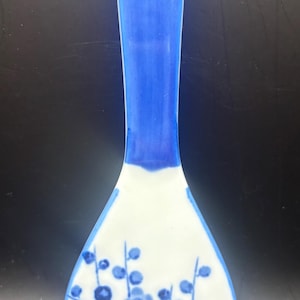 May include: Blue and white ceramic spoon rest with a floral design. The handle is solid blue, transitioning to white with blue floral patterns on the spoon's surface. Features a hole for hanging.