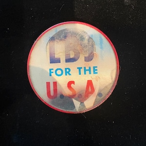 Puede incluir: Un botón redondo con borde rojo y la imagen de un hombre. El botón tiene el texto "LBS FOR THE USA" en azul y rojo. El botón está sobre un fondo negro.