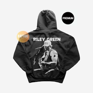 May include: Black hoodie with a white graphic of Riley Green performing, with the text "RILEY GREEN" across the upper back. A "PREMIUM" circle is in the upper right corner. The hoodie has a hood and long sleeves.