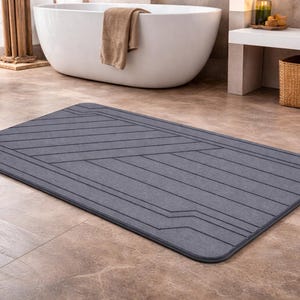 May include: A rectangular gray bath mat with a geometric design of angled and straight lines. The mat is placed on a tiled floor in a bathroom setting, with a white bathtub and wicker baskets in the background.