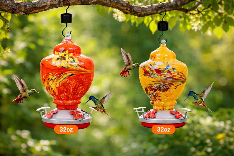 May include: Two 32oz glass hummingbird feeders hanging from a tree branch. One feeder is vibrant orange with yellow and black swirls, and the other is bright yellow with red and blue streaks. Both feature red flower-shaped feeding ports.