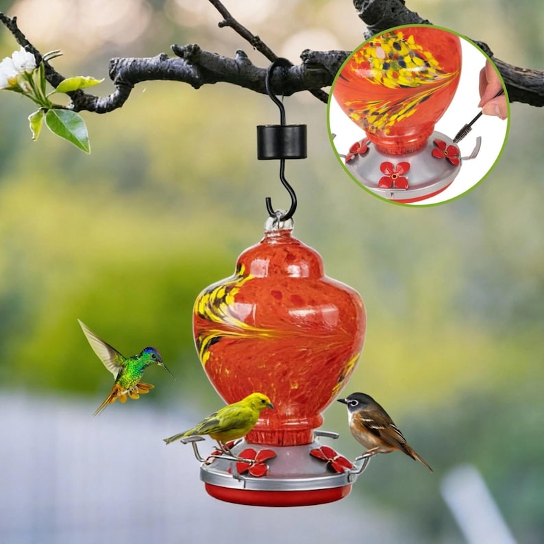 May include: A hummingbird feeder featuring a hand-blown glass reservoir with a swirling orange and yellow pattern. It has a silver metal base with red flower-shaped feeding ports and perches, suspended by a black metal hook from a tree branch.