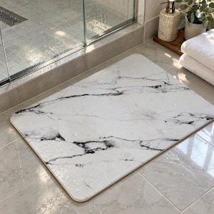 May include: A rectangular bath mat featuring a white surface with black marble veining patterns. The mat has rounded corners and a smooth, flat texture with several small water droplets scattered across the top surface.