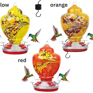 May include: Three glass hummingbird feeders with metal bases and red flower-shaped feeding ports. The feeders are labeled yellow, orange, and red, featuring swirling multi-colored patterns in the glass reservoirs and black metal hanging hooks.