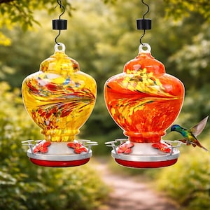 May include: Two glass hummingbird feeders hanging from black metal hooks. One feeder is yellow with red and orange swirls, and the other is orange with yellow and red swirls. Both have silver bases with red flower-shaped feeding ports.