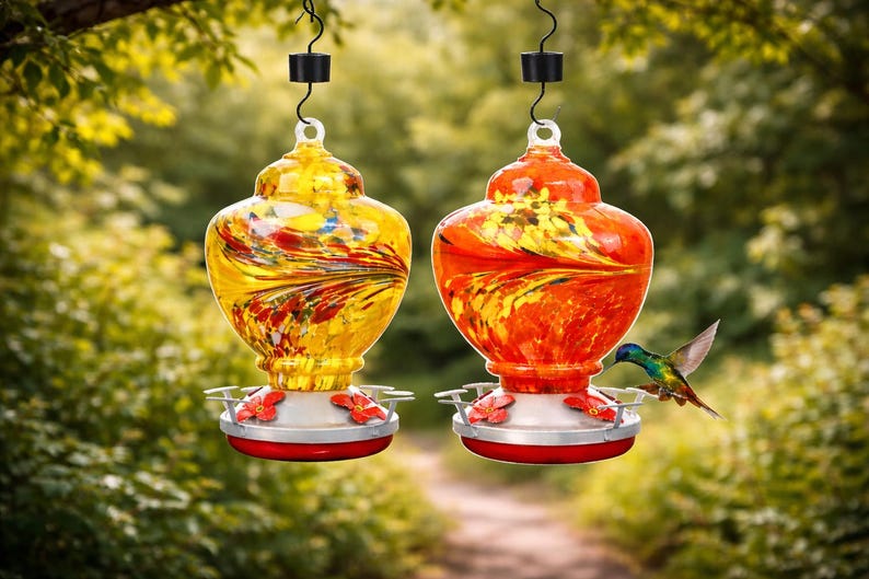May include: Two glass hummingbird feeders hanging from black metal hooks. One feeder is yellow with red and orange swirls, and the other is orange with yellow and red swirls. Both have silver bases with red flower-shaped feeding ports.