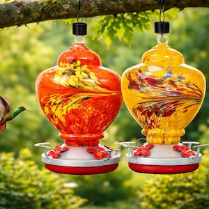May include: Two glass hummingbird feeders hanging from a tree branch. One feeder is vibrant orange with yellow swirls, and the other is bright yellow with orange and red streaks. Both feature red flower-shaped feeding ports and a red base.