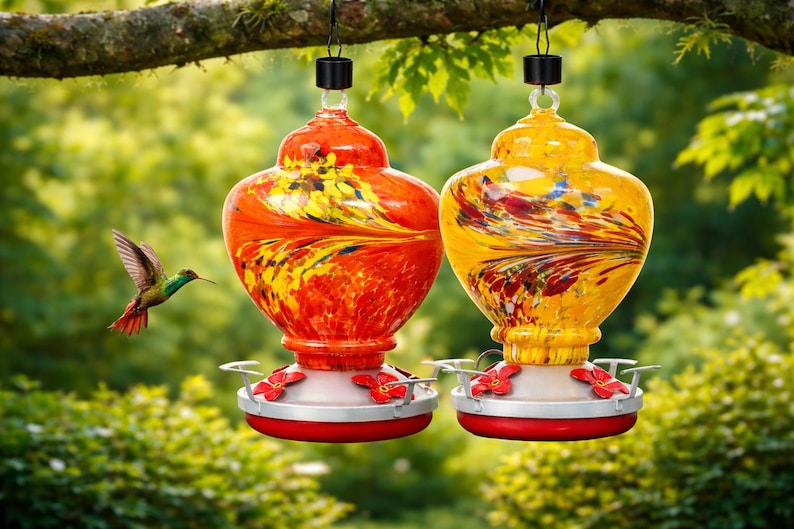 May include: Two glass hummingbird feeders hanging from a tree branch. One feeder is vibrant orange with yellow swirls, and the other is bright yellow with orange and red streaks. Both feature red flower-shaped feeding ports and a red base.