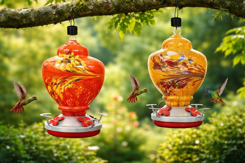May include: Two glass hummingbird feeders hanging from a tree branch. One feeder is bright orange with yellow swirls, and the other is golden yellow with dark red streaks. Both have silver bases with red flower-shaped feeding ports.