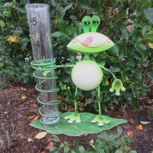 May include: A whimsical green metal frog rain gauge. The frog has a white belly, a green head with large eyes, and a pink tongue. The gauge is clear with measurements in inches, held by a green coil.