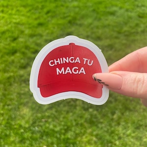 Può includere: Un adesivo a forma di cappellino da baseball rosso con la scritta bianca "CHINGA TU MAGA". L'adesivo è tenuto su uno sfondo erboso verde.