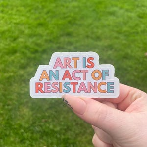 May include: A colorful sticker with the text "ART IS AN ACT OF RESISTANCE" in a variety of bright colors. The sticker is held against a backdrop of green grass.