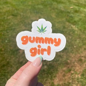 Puede incluir: Una pegatina blanca con las palabras "gummy girl" en naranja, contorneadas en blanco. Una hoja de cannabis verde está en la parte superior. La pegatina se sostiene contra un fondo de hierba verde.