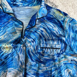 May include: Blue pajama top featuring a swirling, impressionistic design in shades of blue, white, and yellow, reminiscent of Van Gogh's "Starry Night." The top has black trim, a chest pocket, and button closures. The fabric appears to be silky.