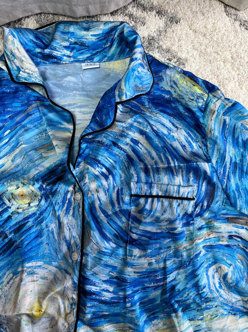 May include: Blue pajama top featuring a swirling, impressionistic design in shades of blue, white, and yellow, reminiscent of Van Gogh's "Starry Night." The top has black trim, a chest pocket, and button closures. The fabric appears to be silky.