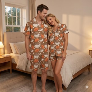 May include: Matching brown pyjama sets with a coffee and croissant pattern. The sets include a short-sleeved button-down shirt and shorts. The pyjamas are worn by a man and a woman standing in a bedroom.