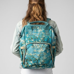 May include: A teal backpack featuring a floral design reminiscent of Van Gogh's Almond Blossoms. The backpack has multiple compartments, a front pocket, and adjustable straps. It is being worn by a person.