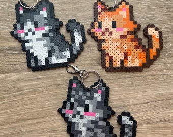May include: Three pixel art cat keychains in gray, orange, and black. Each cat has a pink nose and is attached to a silver keychain. The keychains are made of small, colorful beads and are sitting on a wooden surface.