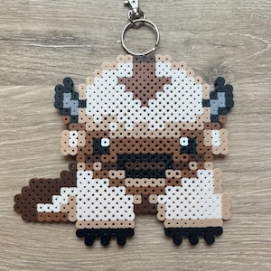 May include: A handmade keychain featuring a pixelated design of Appa from Avatar: The Last Airbender. The keychain is made from fused plastic beads in shades of brown, tan, white, and black. It includes a silver-colored metal keyring and clasp.
