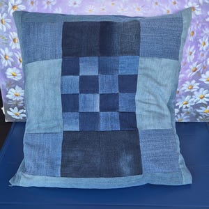 Peut inclure: Un coussin carré fabriqué à partir de différentes nuances de tissu denim bleu. Le panneau central présente un motif en damier tissé de carrés de denim bleu foncé et moyen, flanqué de panneaux de denim unis dans des tons bleu clair et foncé.