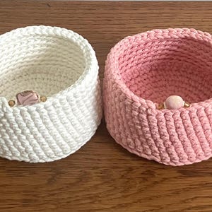 Op de afbeelding: Twee handgemaakte gehaakte manden. Eén is wit, de andere roze. Elke mand heeft een decoratieve kraal aan de voorkant. De manden zijn rond met een licht uitlopende vorm, perfect voor opslag of decoratie.