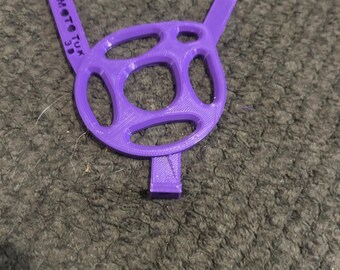 Op de afbeelding: Een paars 3D-geprint MotoTuk 3D-apparaat. Het apparaat heeft een cirkelvormig ontwerp met meerdere ovale uitsparingen en twee verlengde armen. De tekst "MotoTuk 3D" is op een van de armen gedrukt.