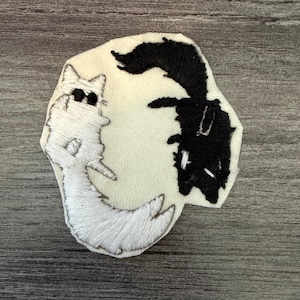 May include: Embroidered patch featuring a white cat and a black dog in a yin-yang design. The cat is white with black eyes and whiskers, while the dog is black. The patch is on a cream-colored background and has a stitched border.