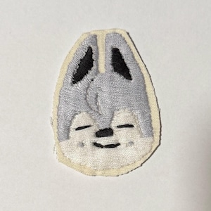 May include: Embroidered patch featuring a stylized wolf face with closed eyes. The patch has a light gray face, black ears, and a black nose and mouth. The patch is outlined with a cream-colored border.