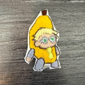 May include: Embroidered patch featuring a cartoon character in a banana costume. The patch has a yellow banana peel, a yellow outfit, and gray pants. The character has blonde hair, green eyes, and wears round glasses. The patch is on a gray wooden surface.