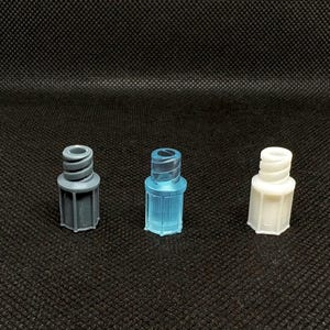 May include: Three plastic connectors in shades of gray, blue, and white are arranged on a black surface. Each connector has a cylindrical body with a ribbed top and a hexagonal base. The connectors are designed for connecting tubes or hoses.