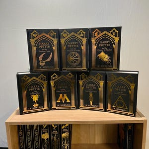 May include: A collection of seven hardcover books from the Harry Potter series, displayed on a wooden shelf. The books have black covers with gold lettering and illustrations. The titles include "Sorcerer's Stone," "Chamber of Secrets," and "Prisoner of Azkaban."