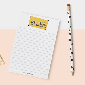 May include: A white lined notepad featuring a yellow rectangular graphic with the blue text BELIEVE at the top and the text JONES STREET PRESS at the bottom, accompanied by a white pencil with black polka dots and a gold paper clip.