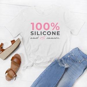 May include: White t-shirt with pink text that reads "100% Silicone and 0% cancer". The shirt is laid out on a white wooden surface with a pair of brown sandals and blue jeans.