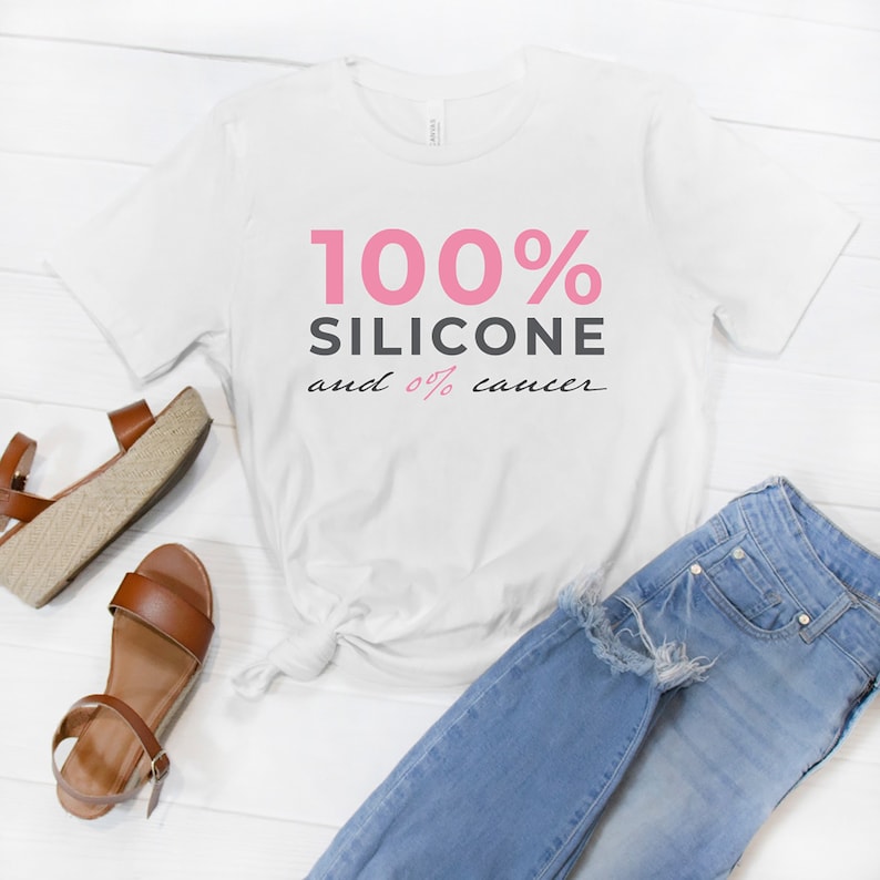Pode incluir: Camiseta branca com texto rosa que diz "100% Silicone e 0% c&acirc;ncer". A camiseta est&aacute; colocada sobre uma superf&iacute;cie de madeira branca com um par de sand&aacute;lias marrons e uma cal&ccedil;a jeans azul.