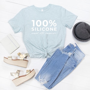 Pode incluir: Uma camiseta azul claro com o texto "100% Silicone e 0% C&acirc;ncer" impresso em branco. A camiseta est&aacute; colocada sobre uma superf&iacute;cie de madeira branca com um par de sand&aacute;lias douradas, cal&ccedil;as de ganga azuis e um chap&eacute;u preto.