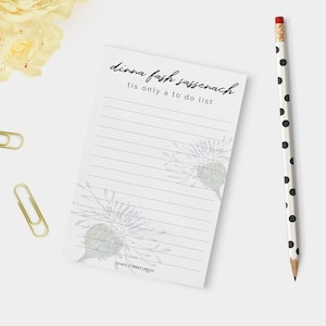 Pode incluir: Um bloco de notas pautado com o texto "dinna fash sassenach tis only a to do list" e ilustrações florais. Um lápis de bolinhas preto e branco e clipes de papel dourados também estão na imagem.