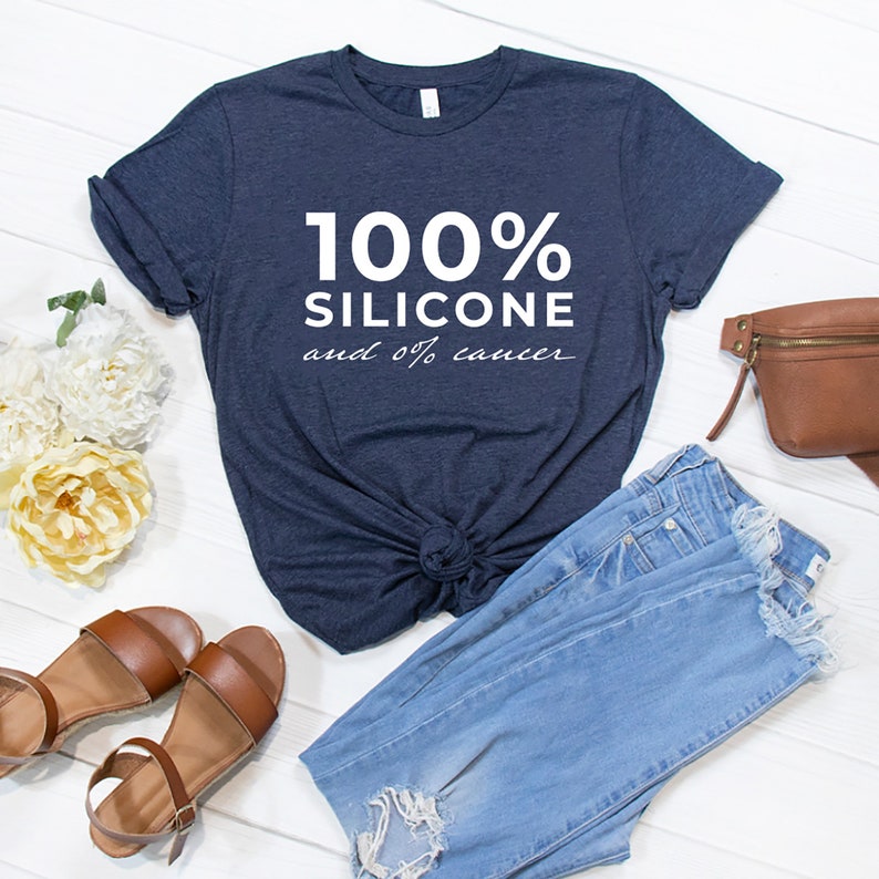 Pode incluir: Uma camiseta azul marinho com o texto "100% Silicone e 0% c&acirc;ncer" em branco. A camiseta est&aacute; colocada sobre uma superf&iacute;cie de madeira branca com um par de sand&aacute;lias marrons, cal&ccedil;as de ganga azuis e uma pochete de couro marrom.