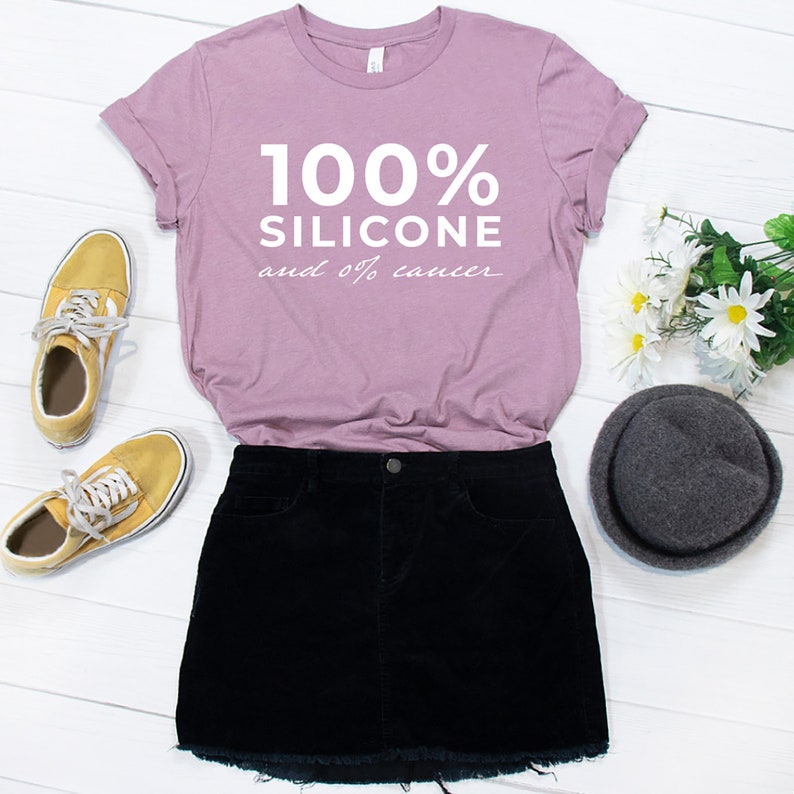 Pode incluir: Uma camiseta de cor lil&aacute;s claro com o texto "100% SILICONE e 0% cancro" em branco. A camiseta &eacute; combinada com uma minissaia preta, t&ecirc;nis amarelos e um chap&eacute;u cinza.