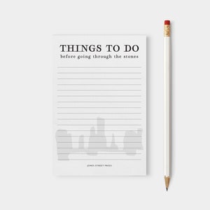 Pode incluir: Um bloco de notas branco com o texto "THINGS TO DO before going through the stones" e páginas pautadas. Um lápis branco com uma borracha vermelha está posicionado à direita do bloco de notas. O bloco de notas tem um design de aquarela cinza na parte inferior.