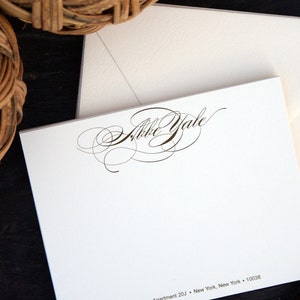 Puede incluir: Juego de papelería blanco con una tarjeta con el nombre "Abbe Yale" en escritura elegante, acompañada de un sobre a juego. La papelería se encuentra sobre un fondo oscuro con una cesta tejida en la esquina superior izquierda.