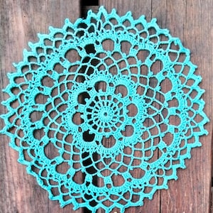 May include: A round, turquoise crocheted doily. The doily features a detailed openwork design with a central spiral and a scalloped edge. The handmade piece is displayed on a rustic wooden surface, showcasing its intricate texture and color.