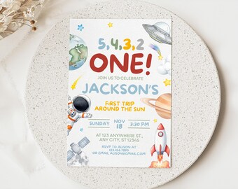Könnte beinhalten: Eine Geburtstagseinladung zum Thema Weltraum mit dem Text "5,4,3,2 ONE!" und "Jackson's First Trip Around The Sun." Das Design umfasst Illustrationen eines Planeten, eines Astronauten, einer Rakete und eines UFOs. Die Einladung hat einen weißen Hintergrund.