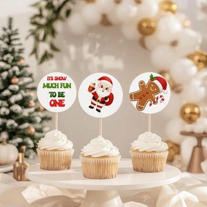 May include: Three cupcakes with white frosting on a white cake stand. Each cupcake has a decorative topper with a Christmas theme. One topper reads "It's snow much fun to be one". The other toppers feature Santa Claus and a gingerbread man.