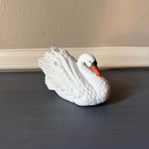May include: A white ceramic swan figurine with detailed feather textures. The swan has a black beak and orange accents. The figurine is resting on a dark gray surface, with a neutral-toned wall in the background.