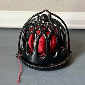 May include: A black, ornate yarn holder with a red ball of yarn inside. The holder has a cage-like design with a handle on top. The yarn is visible through the openings in the holder. The holder sits on a gray surface.