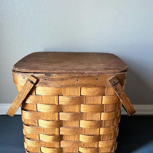 May include: A woven rectangular picnic basket with a wooden lid and two wooden handles. The basket is made of light brown woven material. The lid is a darker brown color. The basket is empty.