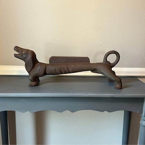 May include: A cast iron dachshund dog doorstop in a brown finish. The dog is in a lying down position with its mouth open and tail curled. The doorstop is on a gray table.