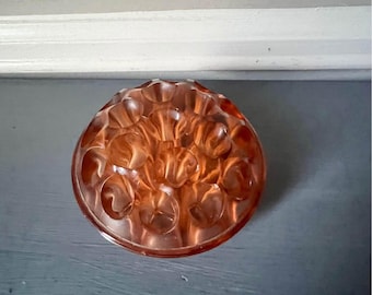 May include: A vintage, round, peach-colored glass flower frog with a textured surface. The flower arranging tool has multiple holes for holding stems. The decorative piece is likely from the mid-century era.