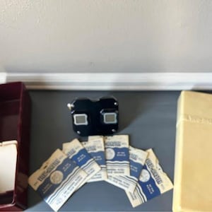 May include: A black plastic View-Master stereoscope viewer accompanied by a collection of paper View-Master reels in their original sleeves, featuring blue and white branding, alongside a maroon storage box and a light-colored rectangular case.