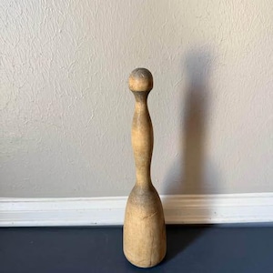 May include: A tall, light-colored wooden kitchen tool with a rounded top, a slender neck, and a wide, heavy base. The wood shows natural grain patterns, variations in tone, and signs of wear, including small surface indentations and marks.